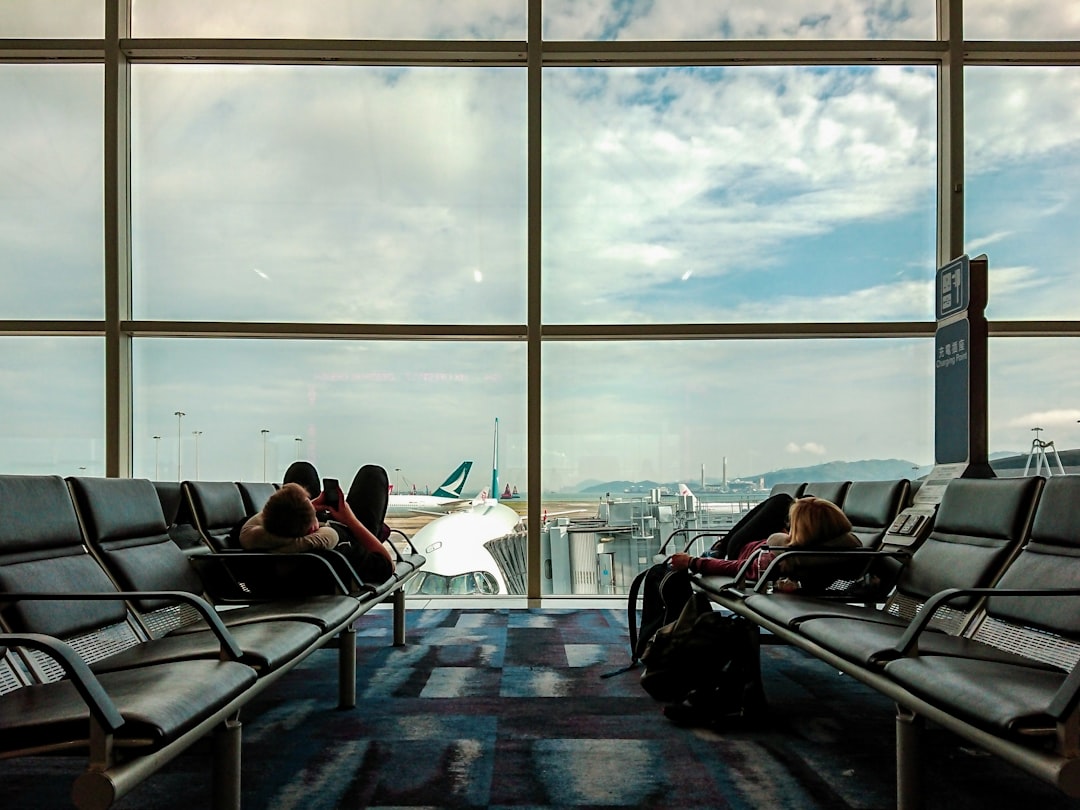 Airport terminal lounge with passengers seated by floor-to-ceiling windows overlooking planes and runway