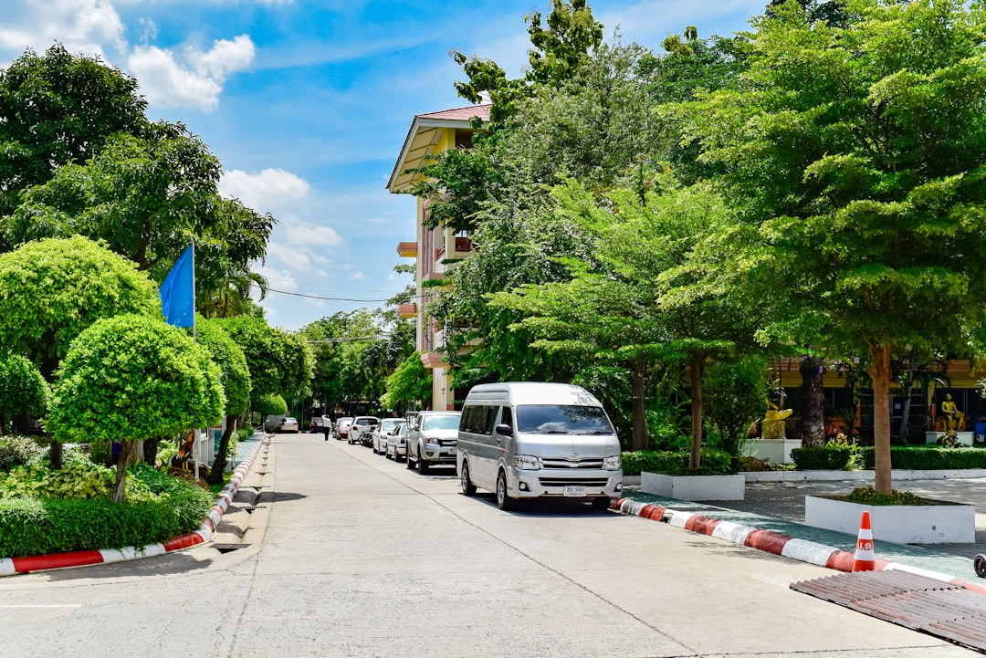 Peaceful Thai street lined with lush green trees, parked vehicles, and a multi-story residential building under blue sky